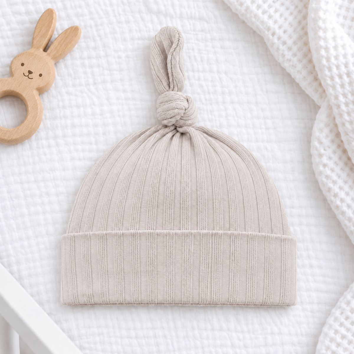 Beige ribbed cotton newborn baby hat with knotted top and folded cuff, laid on white bedding beside a wooden bunny rattle.