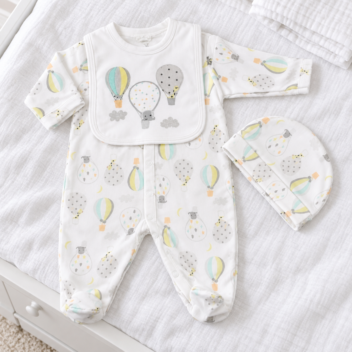 Three-piece white cotton baby sleepsuit set with pastel hot air balloon print, matching baby hat and embroidered-style printed bib.