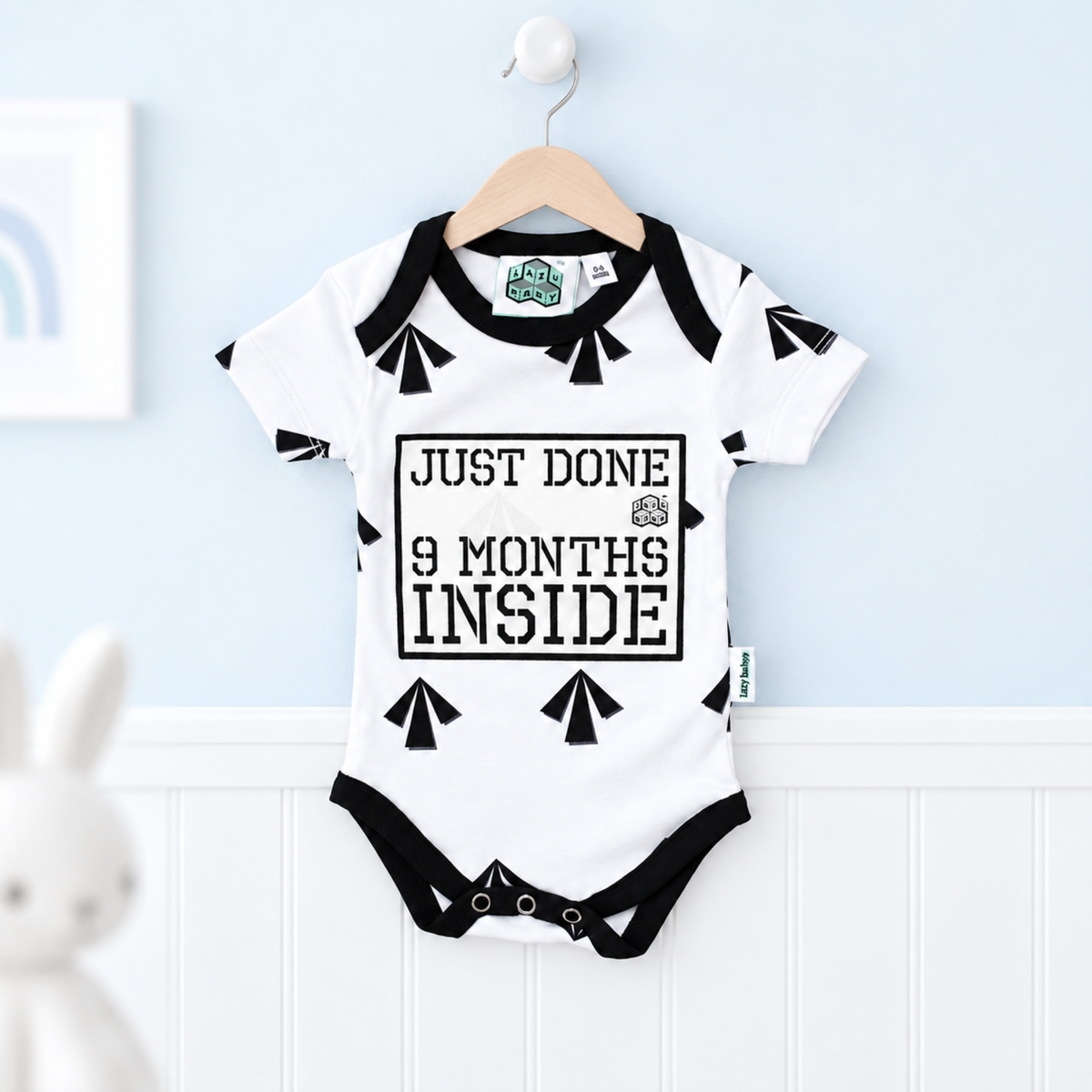 A white baby bodysuit with black trim hangs on a wooden hanger against a light blue wall and white wainscoting. The bodysuit has black graphic shapes and the text 'JUST DONE 9 MONTHS INSIDE' on the front.