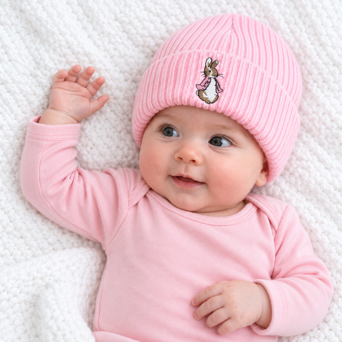Baby girl wearing a pastel pink ribbed knitted hat with an embroidered brown bunny in a pink jacket.