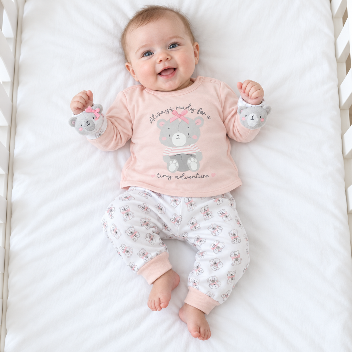 Baby girl wearing pink teddy bear top, matching white bear-print trousers and two grey teddy bear wrist rattles
