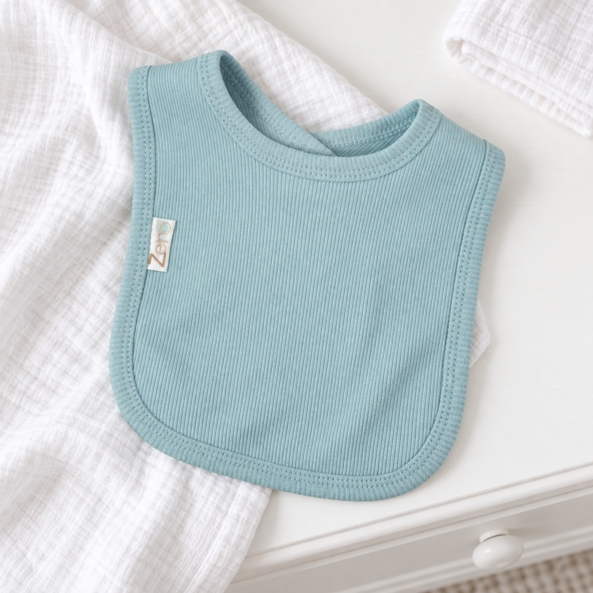 A light blue ribbed fabric baby bib with a small white brand tag, styled on a white wooden surface with white textured muslin cloths.