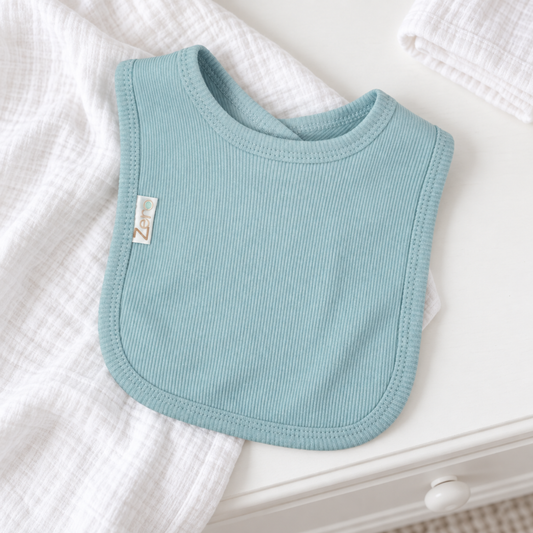 A light blue ribbed fabric baby bib with a small white brand tag, styled on a white wooden surface with white textured muslin cloths.