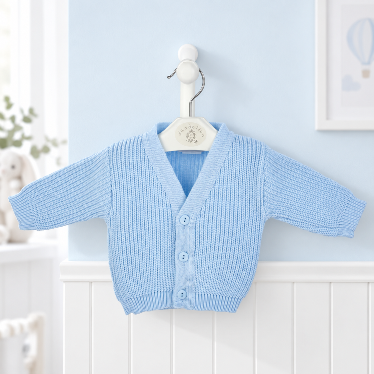 A light blue ribbed knit baby cardigan sweater with long sleeves and three front buttons, hanging on a white hanger against a pale blue wall and white wainscoting.