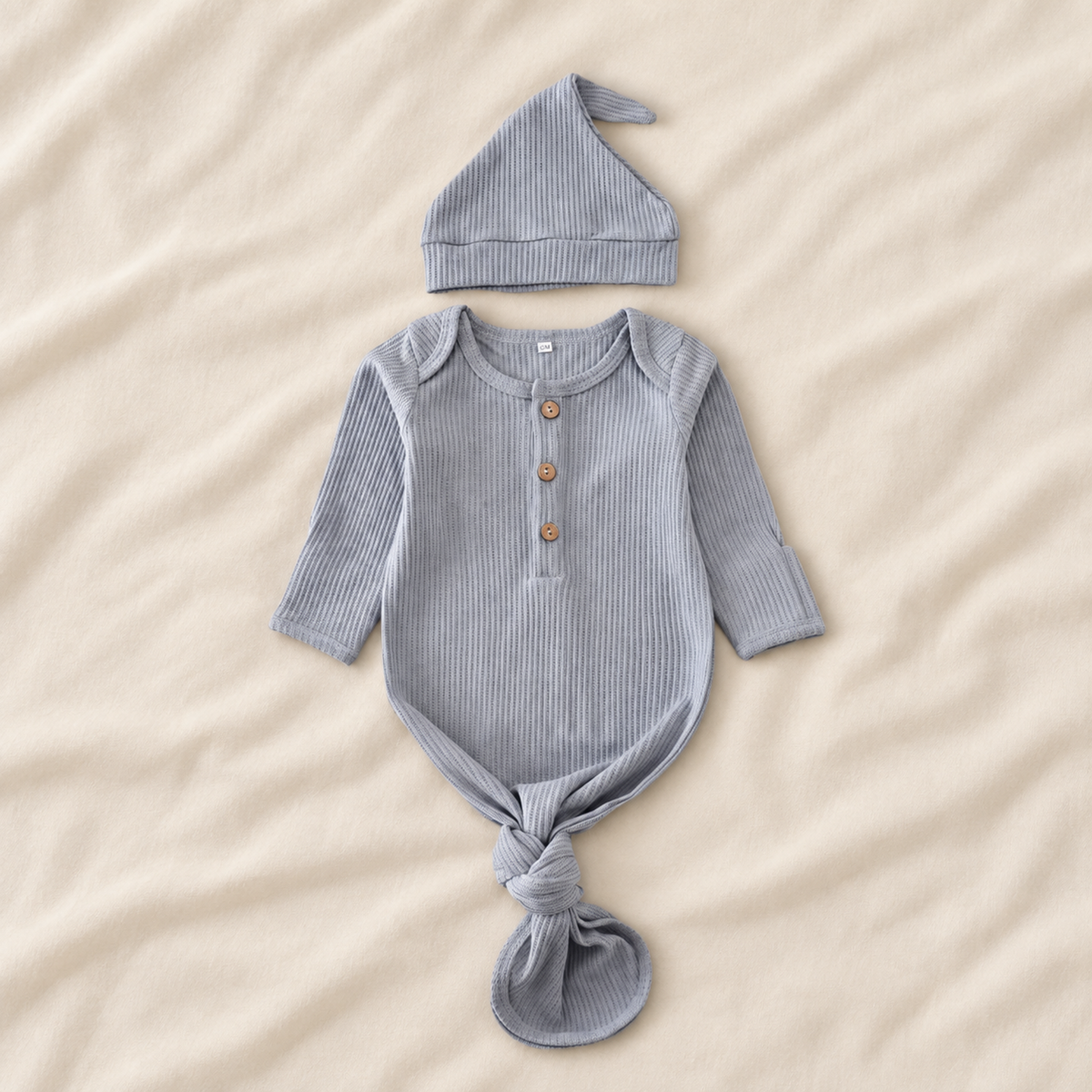 A baby outfit set featuring a long-sleeve ribbed romper and matching hat, laid flat on a beige fabric background. The romper has a buttoned henley neckline and is tied at the bottom, while the matching hat sits above it.