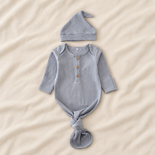 A baby outfit set featuring a long-sleeve ribbed romper and matching hat, laid flat on a beige fabric background. The romper has a buttoned henley neckline and is tied at the bottom, while the matching hat sits above it.