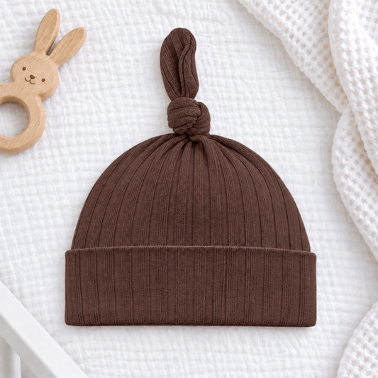 Chocolate brown ribbed cotton baby knot hat with a folded cuff, photographed on white textured bedding beside a wooden bunny teether.