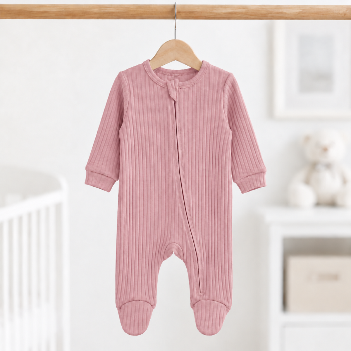 Dusty pink ribbed cotton baby girl sleepsuit with long sleeves, zip fastening and enclosed feet, size 0–3 months, hanging in a nursery.
