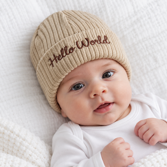 Newborn baby wearing a beige ribbed knitted hat with brown Hello World embroidery on the folded cuff.