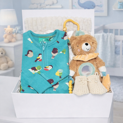 Baby gift set with teal romper, teddy bear, and rattle in a box with a nursery background.
