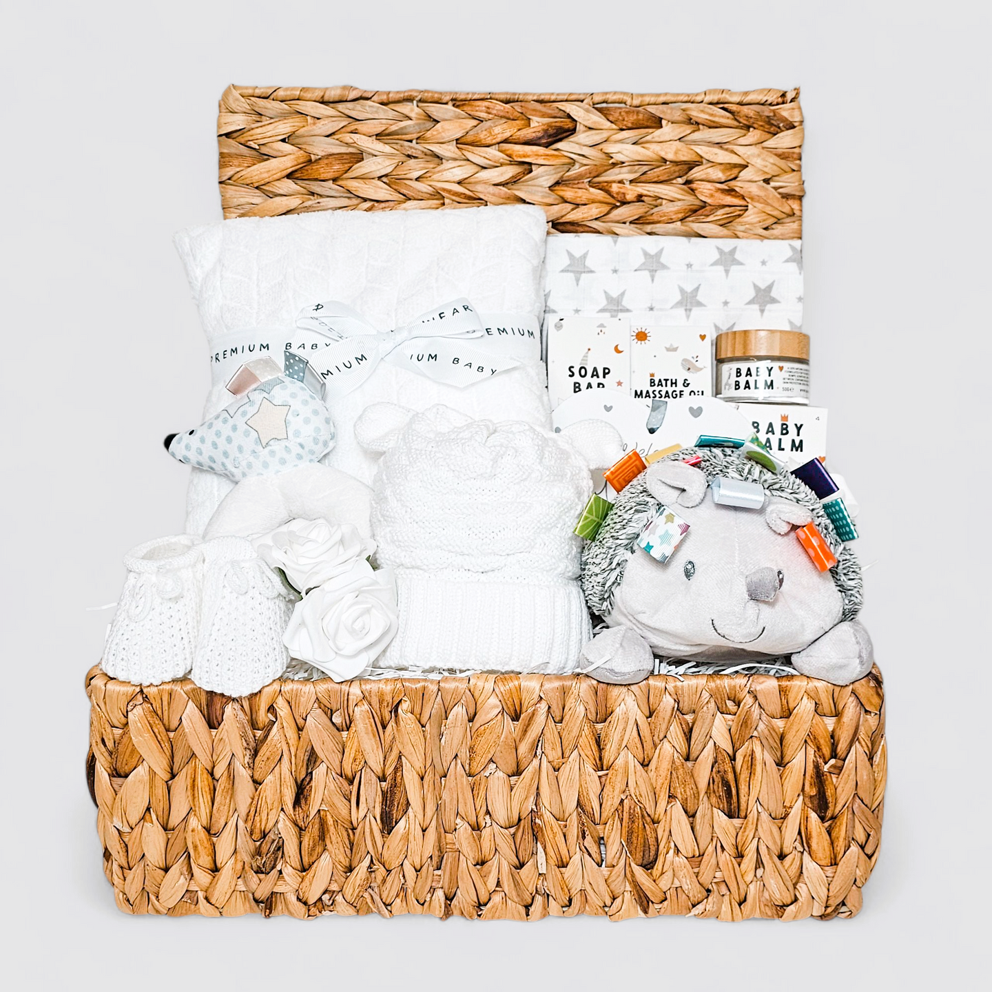 Neutral baby hamper in a basket with white blanket, hedgehog toy, muslin, and baby care items