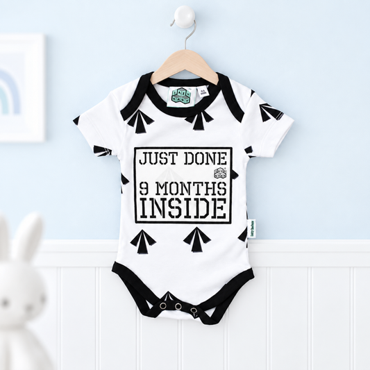 A white baby bodysuit with black trim hangs on a wooden hanger against a light blue wall and white wainscoting. The bodysuit has black graphic shapes and the text 'JUST DONE 9 MONTHS INSIDE' on the front.