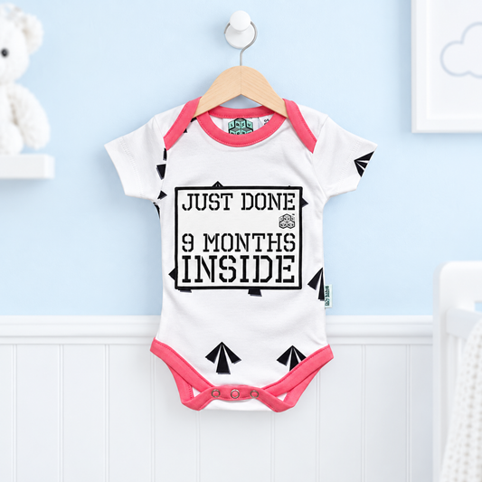 A white baby bodysuit with pink trim and a black graphic reading 'JUST DONE 9 MONTHS INSIDE' hangs on a wooden hanger against a light blue wall with white wainscoting.