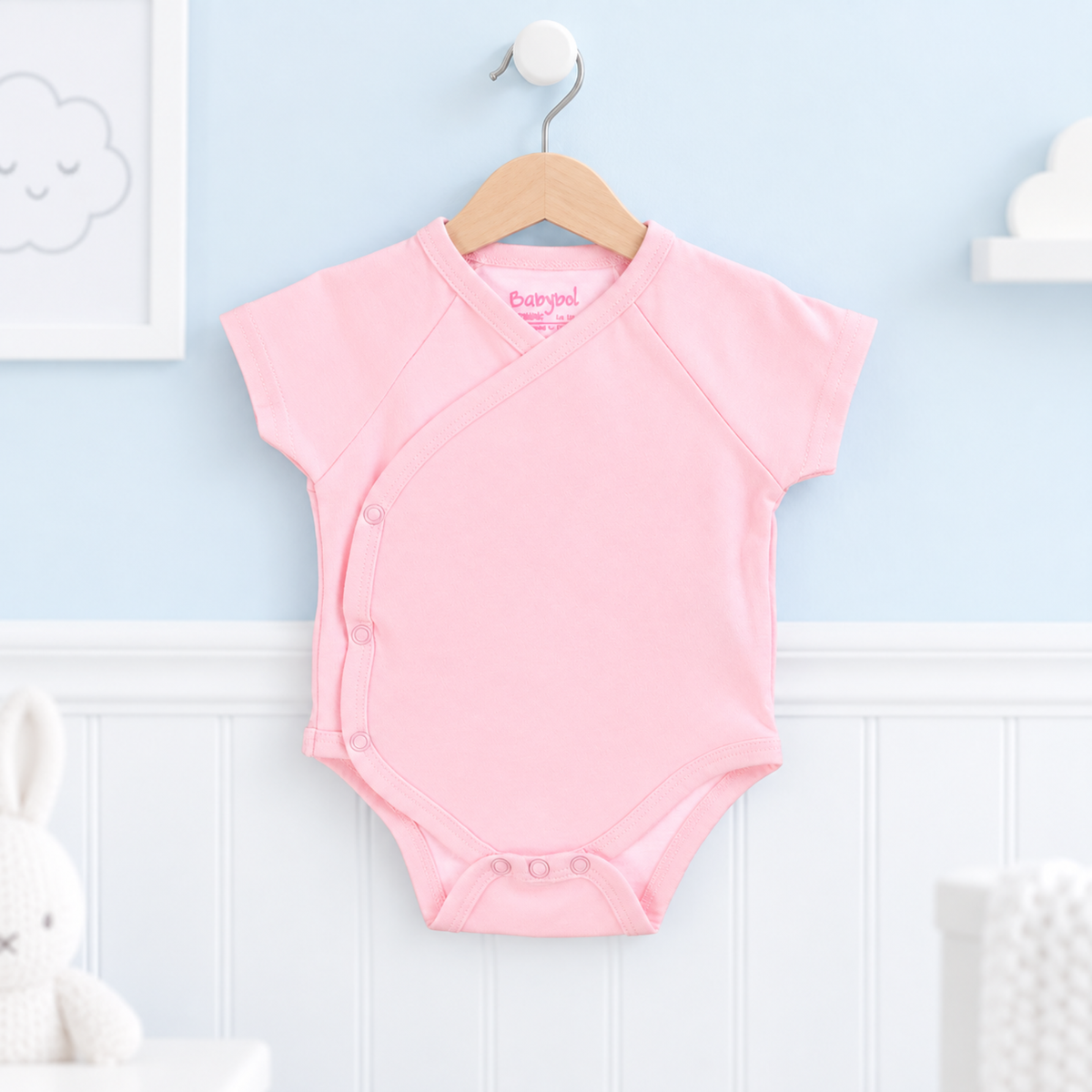 A light pink short-sleeve baby bodysuit with an asymmetrical snap closure, hanging on a wooden hanger against a light blue and white paneled wall.
