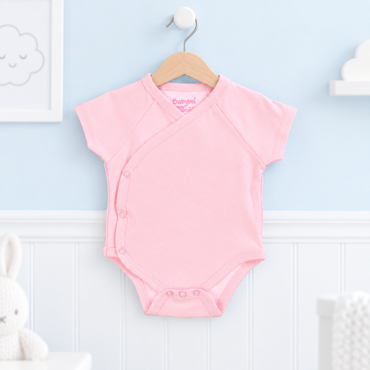 A light pink short-sleeve baby bodysuit with an asymmetrical snap closure, hanging on a wooden hanger against a light blue and white paneled wall.