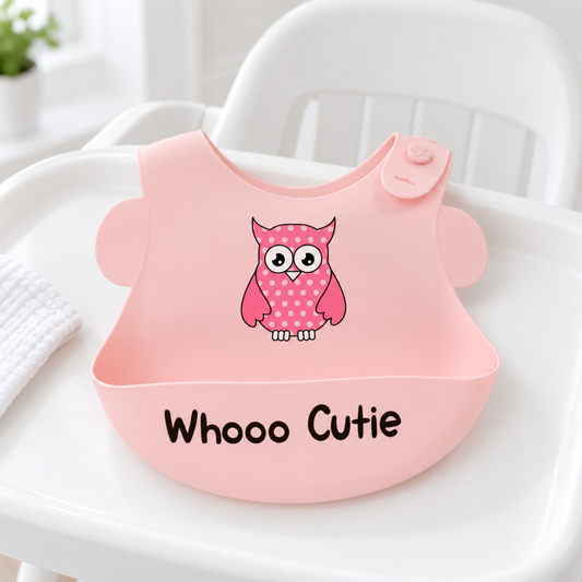 A pink silicone baby bib with a cute pink owl graphic and the text 'Whooo Cutie' printed on the front, displayed on a white high chair tray.