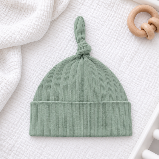 Sage green ribbed cotton baby knot hat with folded cuff, displayed on white bedding beside a natural wooden baby rattle.