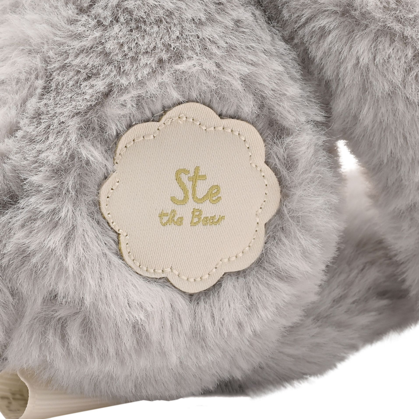 Close-up of a gray faux-fur plush toy with a cream scalloped fabric patch reading 'Ste the Bear' in gold lettering.
