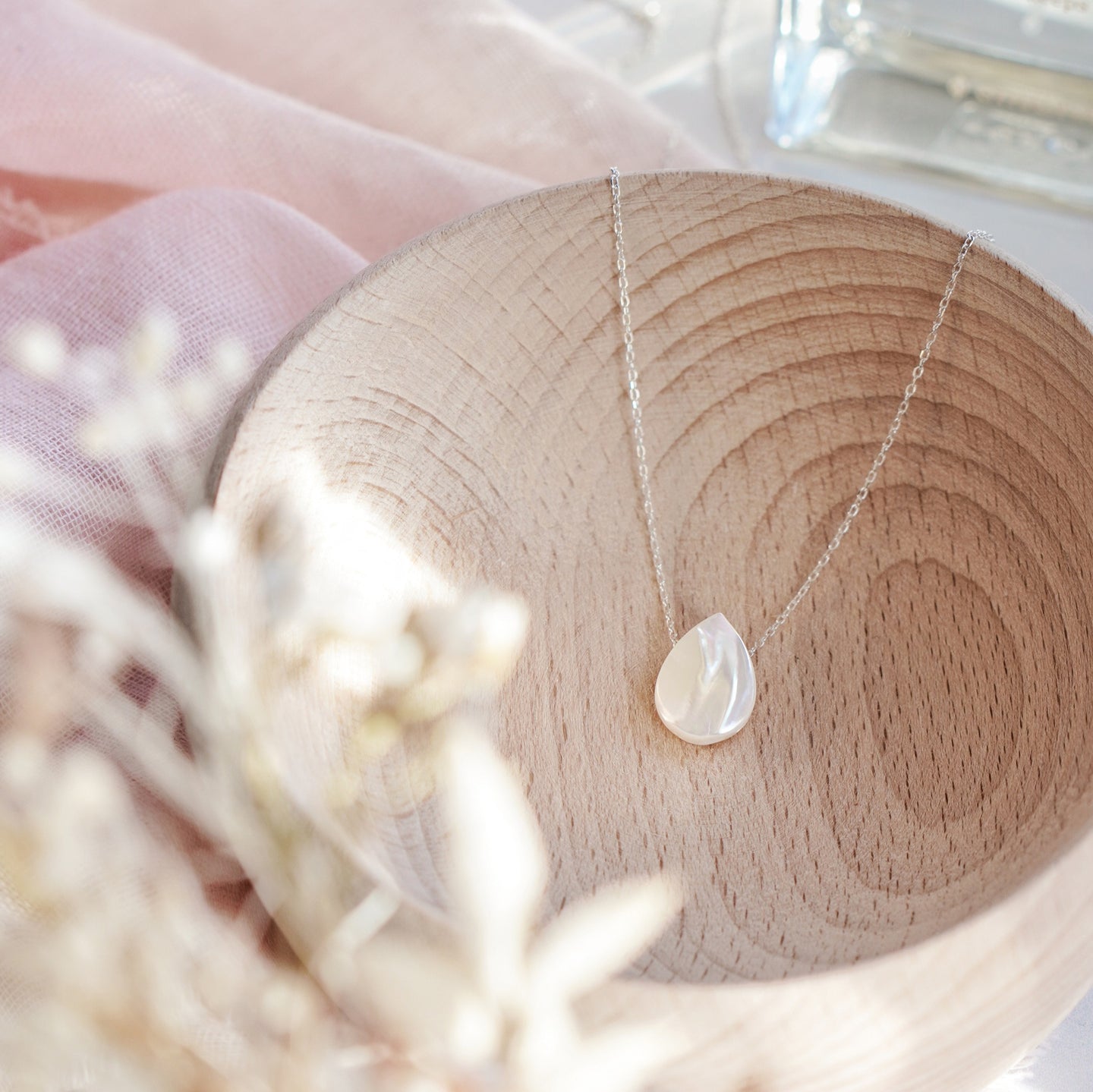 A 925 sterling silver necklace with a white mother of pearl drop pendant, displayed in a bowl, with a blurred background of flowers.